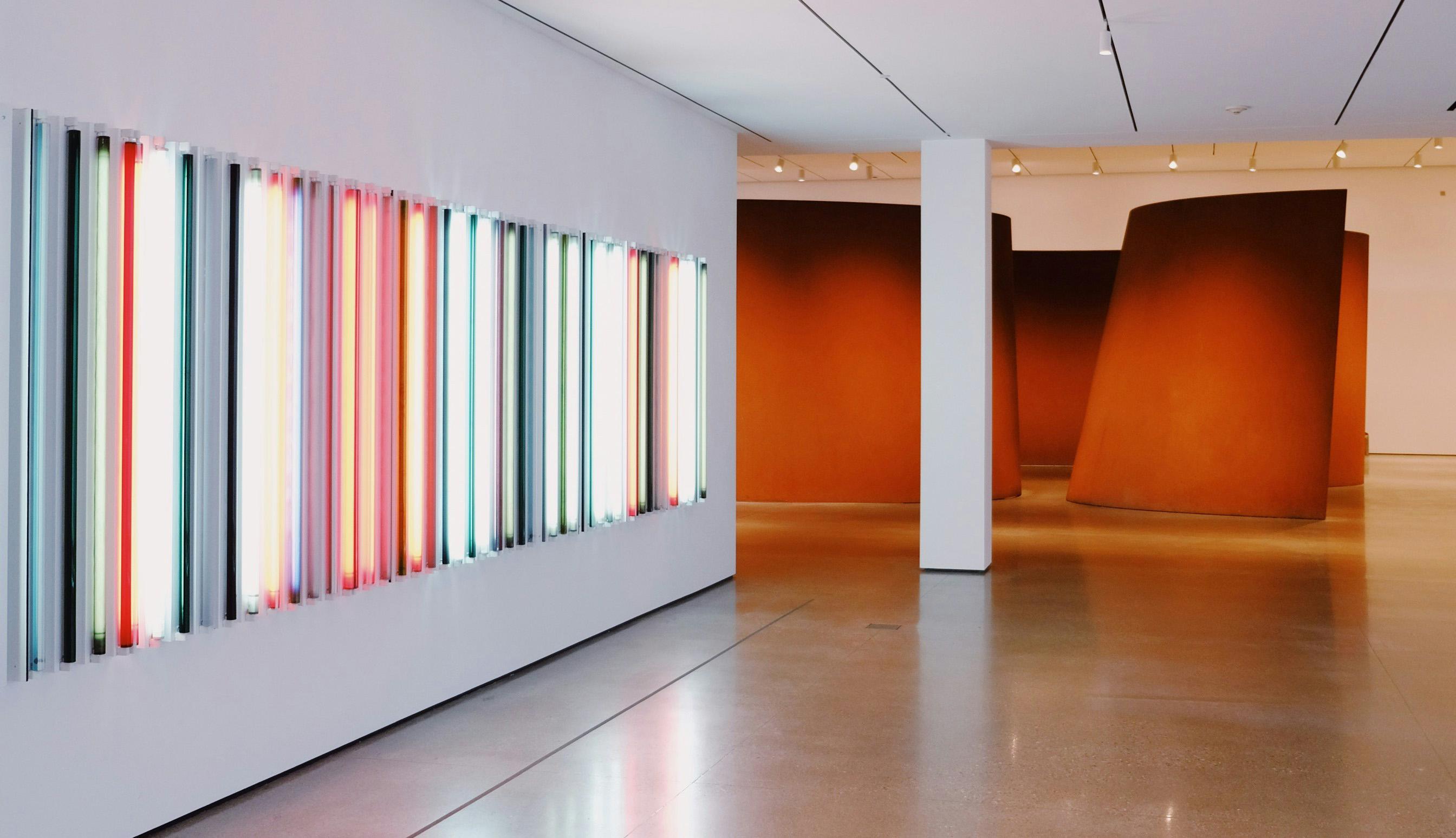 Contemporary gallery interior with a neon light installation and large curved steel sculptures.